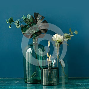 Various artificial flowers and dry plants in transparent vases on the floor against blue wall