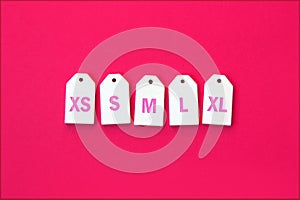 Various clothing sizes XS, S, M, L, XL on white wooden tags on a pink background