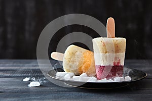 Various coffee, fruit ice cream on a stick are presented in a black plate with slices of ice on a dark wooden background