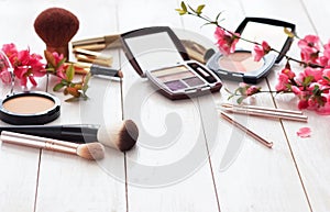 Various cosmetic products for make-up with pink flowers on a white wooden background with copy space.