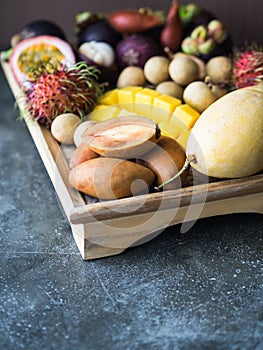 Various fresh Thai fruits - rambutan, mango, mangosteen, longan, sapodilla, passion fruit, salak, on a wooden tray and a gray
