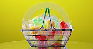 Various fruit candies in transparent packaging lie in a grocery basket on a colorful yellow background