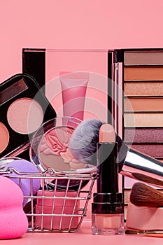 Various make-up products and brushes in shopping cart on pink background. Makeup cosmetics sale concept