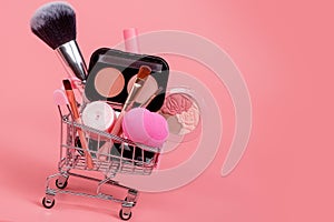 Various make-up products and brushes in shopping cart on pink background. Makeup cosmetics sale concept