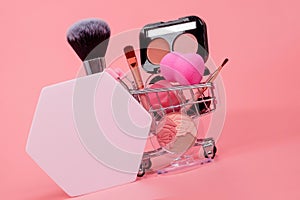 Various make-up products and brushes in shopping cart on pink background. Makeup cosmetics sale concept