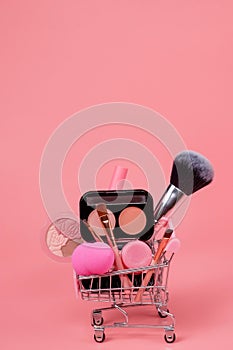 Various make-up products and brushes in shopping cart on pink background. Makeup cosmetics sale concept