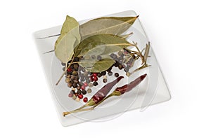 Various peppercorns, dried bay leaves, chili, rosemary on square saucer