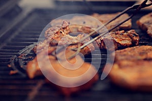 Various pieces of meat are cooked on an iron cast grill for a BBQ - foreground with pliers blanked out blurry for copy space