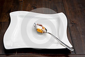 Various pills are on a spoon next to a white plate on a brown table