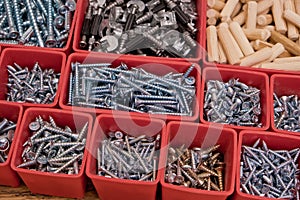 Various size and shape of screws, bolts, wooden pegs selection in red plastic tray box on a wooden background. Assembly kit for fu