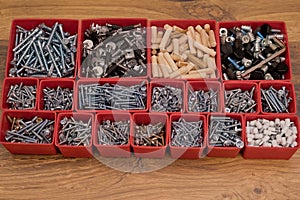 Various size and shape of screws, bolts, wooden pegs selection in red plastic tray box on a wooden background. Assembly kit for fu