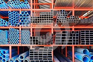 Various sizes of many carbon steel rectangular and square tubes with blue PVC water pipes on storage shelf