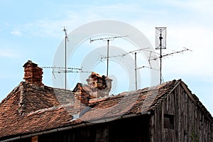 Various sizes TV antennas mounted on multiple metal poles on top of old dilapidated wooden house roof next to destroyed chimneys