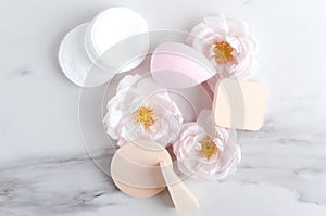 Various types of cosmetic sponges, cotton pads and flowers on the white marble surface