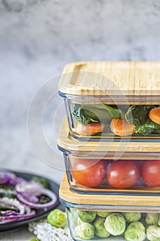 Various vegetables in glass containers: carrots, spinach, tomatoes, Brussels sprouts. Vegan food and snacks in containers, gray