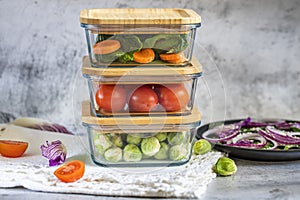 Various vegetables in glass containers: carrots, spinach, tomatoes, Brussels sprouts. Vegan food and snacks in containers, gray