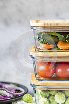 Various vegetables in glass containers: carrots, spinach, tomatoes, Brussels sprouts. Vegan food and snacks in containers, gray