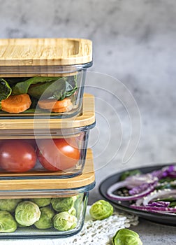 Various vegetables in glass containers: carrots, spinach, tomatoes, Brussels sprouts. Vegan food and snacks in containers, gray