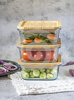 Various vegetables in glass containers: carrots, spinach, tomatoes, Brussels sprouts. Vegan food and snacks in containers, gray