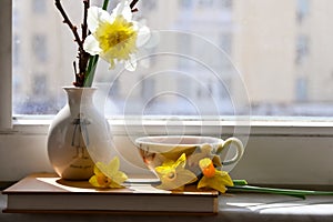 a vase with daffodils , book and a cup of tea are located near the window .