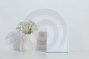 A vase with flowers and a white photo frame on the table. Light stylish background. Preparation for inscriptions, copy space