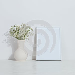 A vase with flowers and a white photo frame on the table. Light stylish background. Preparation for inscriptions, copy space