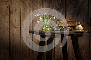 vase with snowdrops, book and other old objects on a vintage stool against a dark rustic wooden wall, spring greeting still life