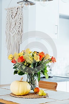 A vase of yellow and orange rose flowers, fresh pumpkin, apple and pear on a kitchen table counter with white modern