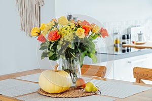 A vase of yellow and orange rose flowers, fresh pumpkin, apple and pear on a kitchen table counter with white modern