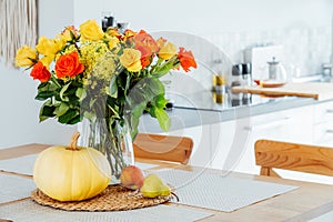 A vase of yellow and orange rose flowers, fresh pumpkin, apple and pear on a kitchen table counter with white modern