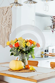 A vase of yellow and orange rose flowers, fresh pumpkin, apple and pear on a kitchen table counter with white modern