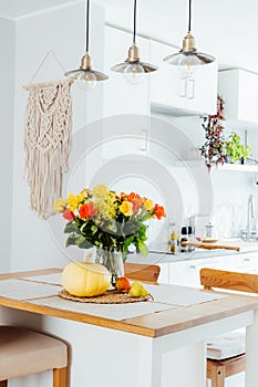 A vase of yellow and orange rose flowers, fresh pumpkin, apple and pear on a kitchen table counter with white modern