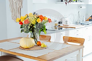 A vase of yellow and orange rose flowers, fresh pumpkin, apple and pear on a kitchen table counter with white modern