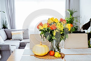 A vase of yellow and orange roses flowers, fresh pumpkin, apple and pear on a kitchen table counter with open space