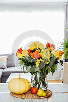 A vase of yellow and orange roses flowers, fresh pumpkin, apple and pear on a kitchen table counter with open space