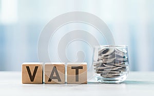 VAT is written on a wooden block beside a clear glass coins jar on a wooden table against a bright white background. Tax and photo