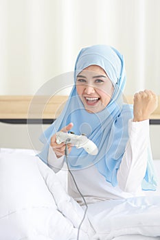 VDO game console station concept. active asian muslim woman wearing white sleepwear sitting on bed, holding joystick and playing