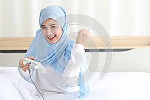 VDO game console station concept. active asian muslim woman wearing white sleepwear sitting on bed, holding joystick and playing