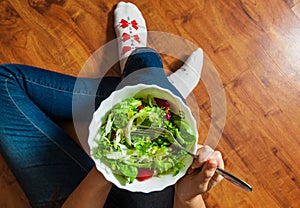 Vegan breakfast meal in bowl with various fresh mix salad leaves and tomato. Girl in jeans holding fork with knees and hands visib