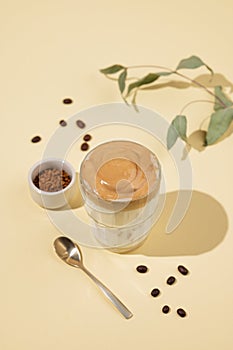 Vegan dalgona coffee. Whipped instant iced coffee with soy milk in a glass on yellow background with coffee bean, spoon and leaves