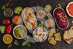 Vegan food background. Vegetarian snacks: hummus, beetroot hummus, green peas dip, vegetables, cereals, tofu. Top view, dark back
