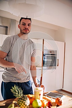 Vegan man in his 20`s has a morning preparation of a smoothie while looking at a recipe on his tablet