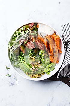 Vegan salad bowl with baked sweet potato, edamame beans, tomatoes, avocado, nuts, tahini dressing and sprouts