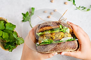 Vegan sandwich with chickpea patty, avocado, cucumber and greens in rye bread in children`s hands