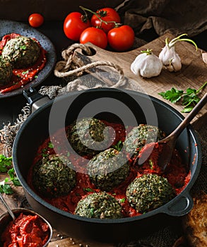 Vegan spinach balls with chickpeas in pan with tomato sauce, onion, parsley on dark background with napkin over wooden table.