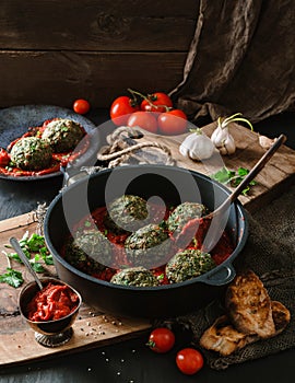 Vegan spinach balls with chickpeas in pan with tomato sauce, onion, parsley on dark background with napkin over wooden table.