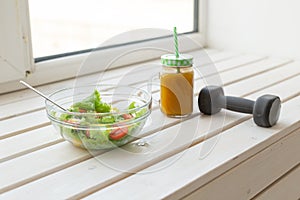 Vegetable salad and fruit smoothies and dumbbell lie on a white windowsill. Concept of healthy lifestyle physical