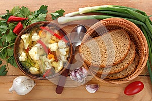 Vegetable soup of cauliflower, carrots, tomato, pepper in a plate with a spoon, bread and onions on a wooden background.