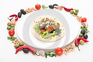 Vegetables arranged in round frame around falafel on pita on plate on white background.