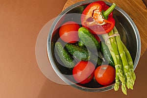 Vegetables lie in a metal bowl: tomatoes, asparagus, cucumbers, red bell peppers. on a wooden board and brown background